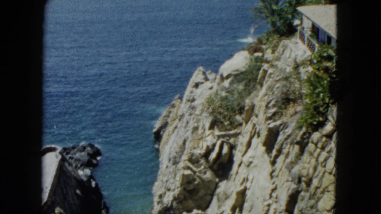 High-angle 8mm view of what appears to be the Acapulco coastline circa 1962, showing cliff face, turquoise Pacific, and a small rooftop structure at the edge.