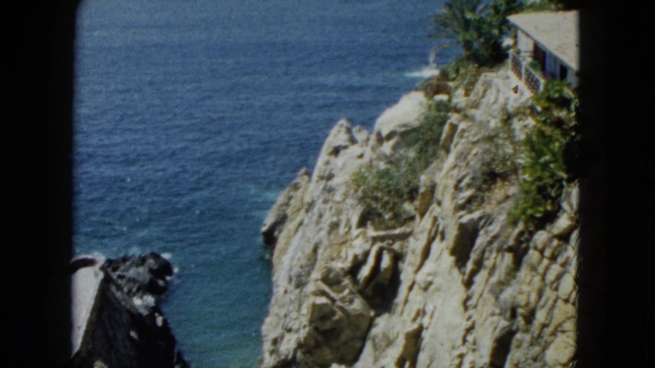 Mid-pan 8mm frame of the Acapulco coastal cliffs circa 1962, showing eroded limestone dropping into turquoise Pacific water and a cave-like rock overhang.
