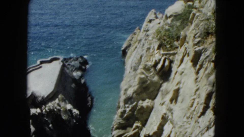 8mm shot circa 1962 of a curved structure on the lower Acapulco cliff face, with dark rock overhang and turquoise Pacific water below.