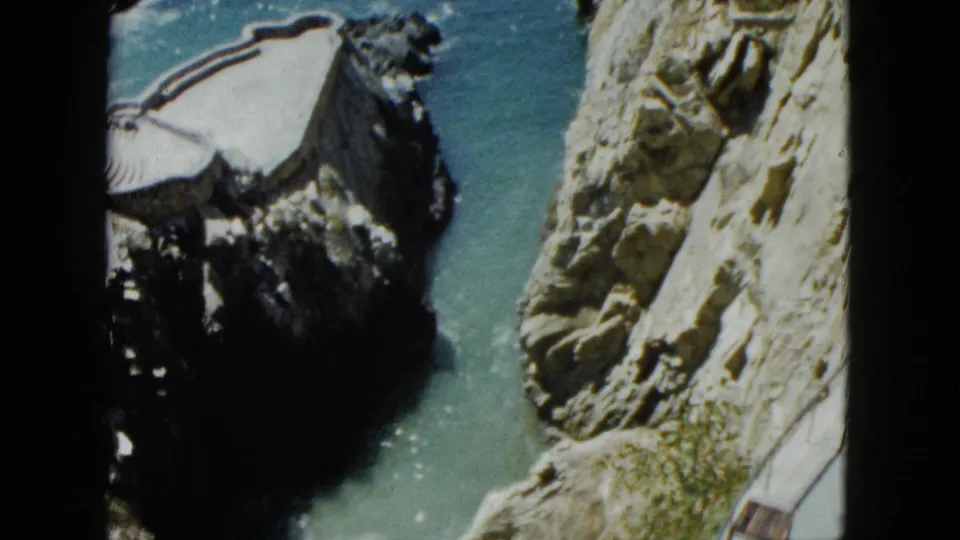 Final pan frame circa 1962 showing a curved structure integrated into the Acapulco cliff above turquoise Pacific water, with rock formations in the foreground.