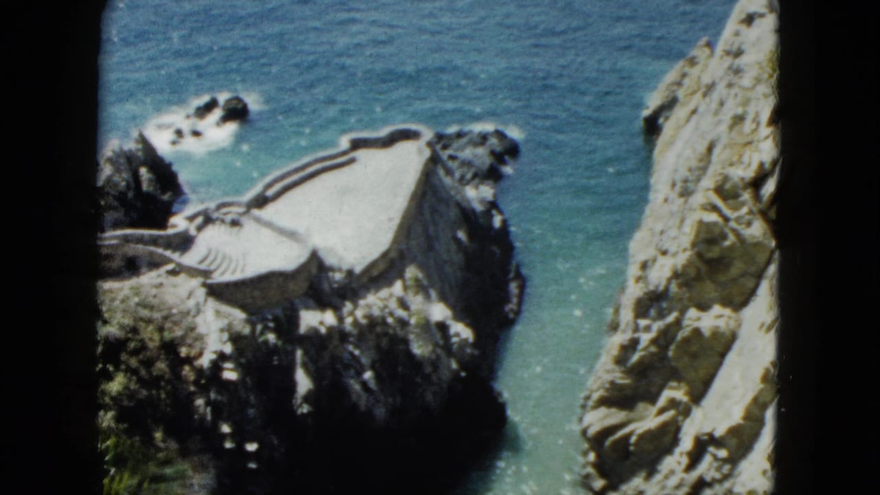 8mm home-movie frame circa 1962, looking down from a cliff at a white structure perched on a rocky outcrop above turquoise Pacific Ocean, Acapulco, Mexico.