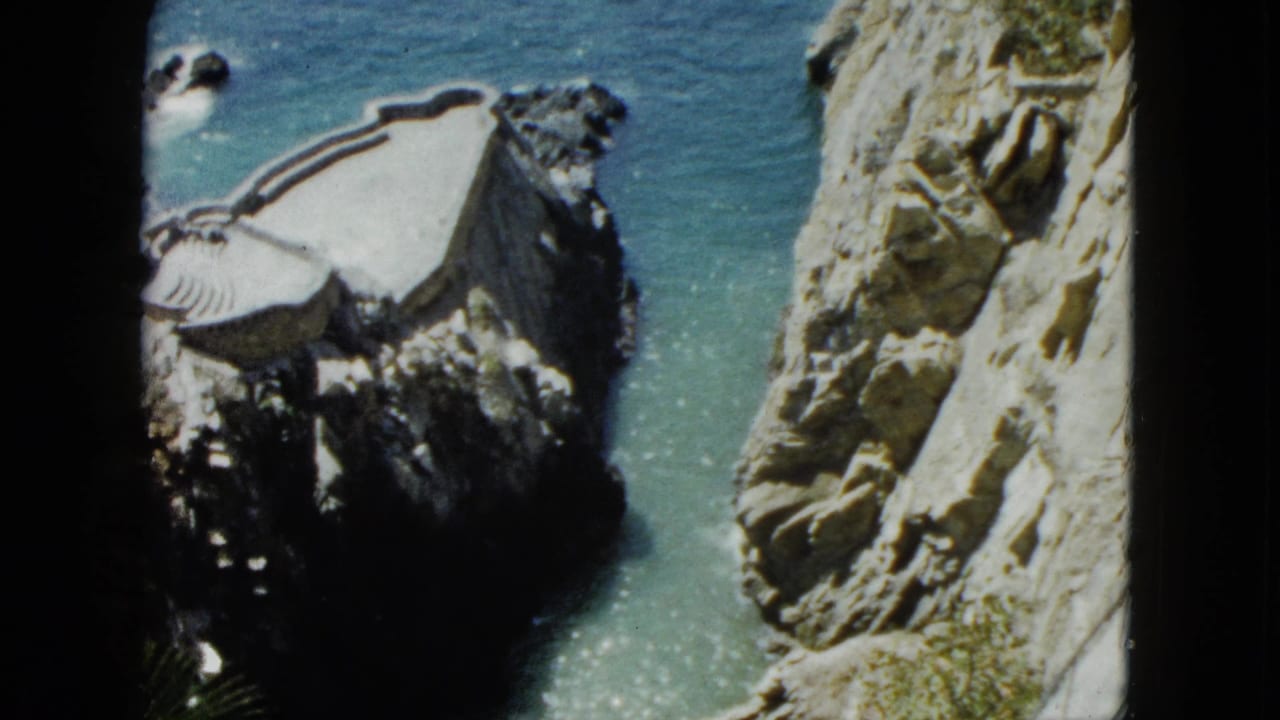 8mm home-movie pan circa 1962, rocky cliff face dominating the frame with white structure at left and turquoise Pacific water above, Acapulco, Mexico.
