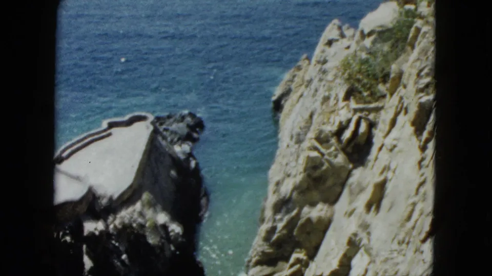 8mm home-movie pan circa 1962, jagged rocky cliffs with white structure at lower left and broad turquoise Pacific water, Acapulco, Mexico.