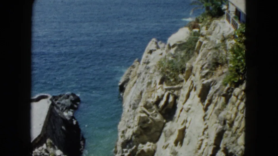8mm home-movie pan circa 1962, rocky cliff face nearly filling frame with sparse scrub at right edge and turquoise Pacific water visible at upper left, Acapulco, Mexico.