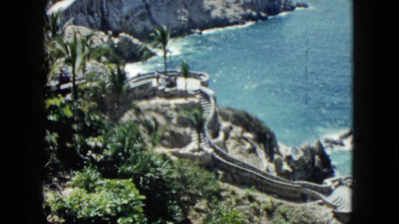 High-angle 8mm view of curved stone staircase and terraced cliff platforms above turquoise sea, palm trees visible, Acapulco, 1962.