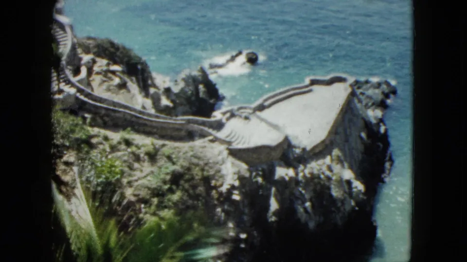 8mm home movie frame showing stone terraced platforms and winding cliff pathway at Acapulco, Mexico, circa 1962; turquoise sea visible at base of cliff with rocky outcroppings.