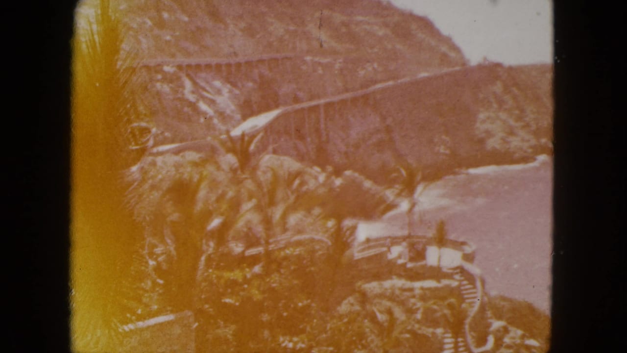 Rocky coastline with palm trees, cliffside bridge or road, and blue sea beyond.