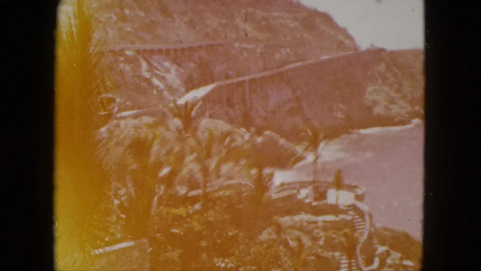 1962 Acapulco coastal view with palms, a stone overlook, rugged cliffs, and a long cliffside bridge or road above the sea.