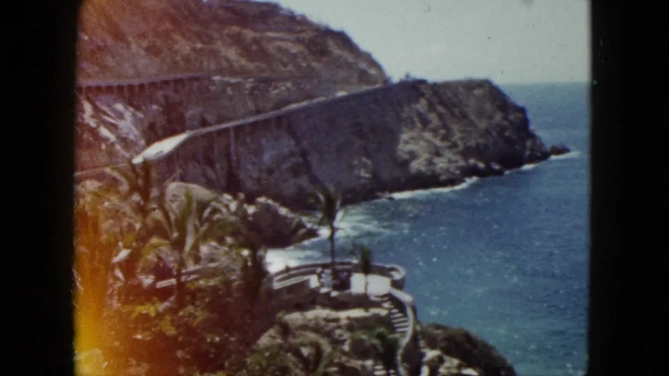Elevated 1962 Acapulco view showing a winding stone staircase, palms, rugged headland, and calm blue water along the rocky shore.