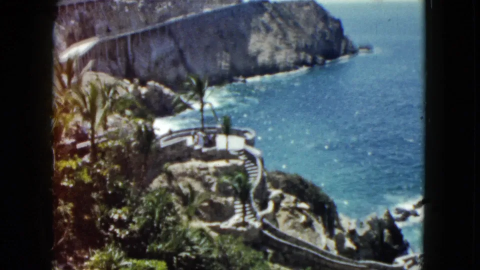 Clear 1962 Acapulco home-movie frame of the rocky coastline, blue sea, palms, winding steps, and cliffside bridge or road in the background.