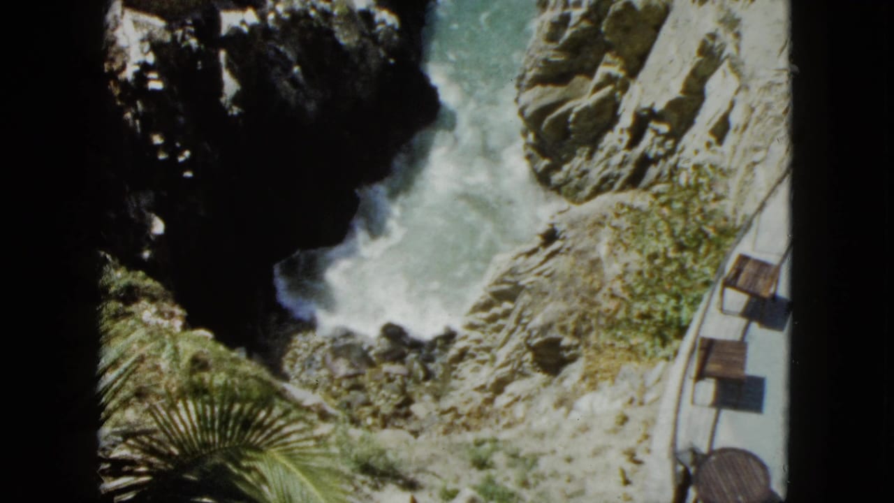 High-angle 8mm shot from 1962 Acapulco showing turquoise waves crashing against rocky cliffs, with a curved clifftop terrace railing, wooden benches, and palm fronds in foreground