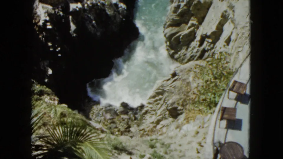 High-angle 8mm shot from 1962 Acapulco showing turquoise waves crashing against rocky cliffs, with a curved clifftop terrace railing, wooden benches, and palm fronds in foreground