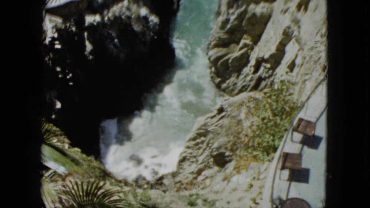 High-angle view of waves crashing against rugged cliffs with a curved terrace walkway and wooden seating visible along the cliff edge