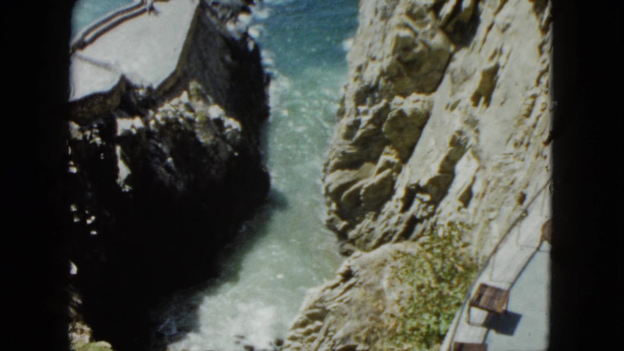 High-angle 8mm view of a rocky coastal chasm with turquoise water and a narrow cliff-edge path with wooden benches.
