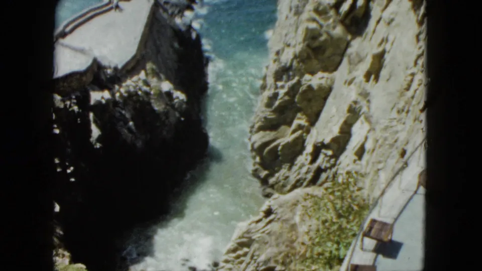 Looking down into a rocky coastal chasm in what appears to be Acapulco, Mexico, circa 1962; turquoise water and a paved tourist path with wooden benches visible.