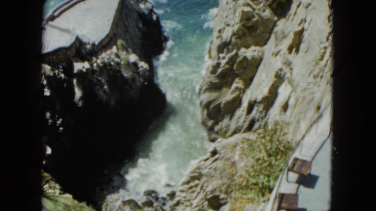 Steady high-angle view of a rocky coastal gorge, likely Acapulco circa 1962; turquoise surf crashes below as wooden benches line a narrow cliff-edge walkway.