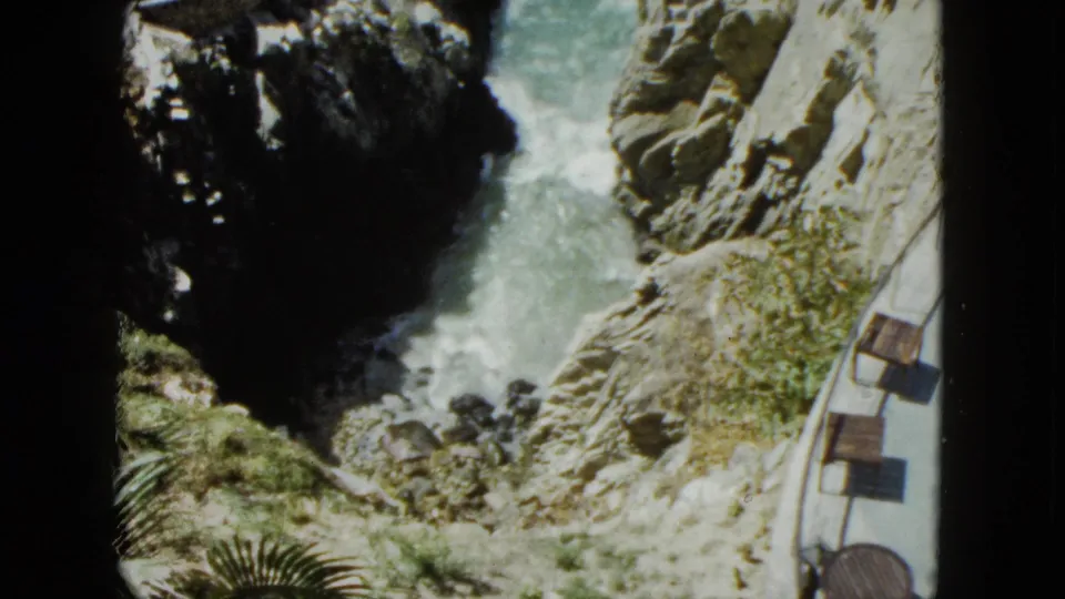 High-angle 8mm view of an Acapulco coastal chasm circa 1962; palm fronds and tropical shrubs appear at the frame edge alongside a curved tourist path with benches.