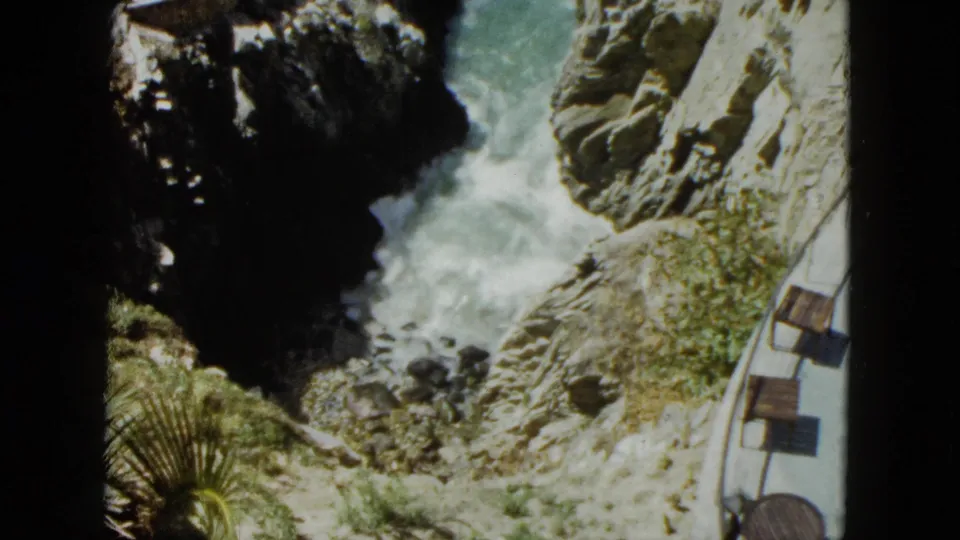 Rocky coastal chasm, likely Acapulco circa 1962; a palm frond enters at lower left as turquoise waves surge below a curved cliff-edge walkway with wooden benches.