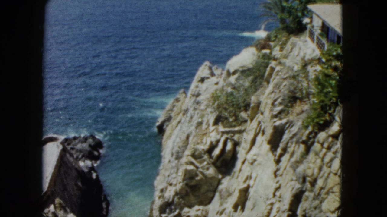 8mm home movie frame from 1962 Acapulco, Mexico: limestone cliff descending to turquoise ocean, cliff-top structure and tropical scrub visible at upper right.