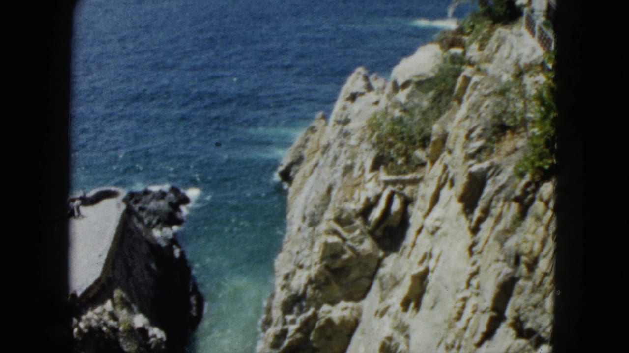 8mm home movie frame from Acapulco, 1962: pan across rugged cliff with deep blue Pacific, small platform or structure at cliff top with railing visible.