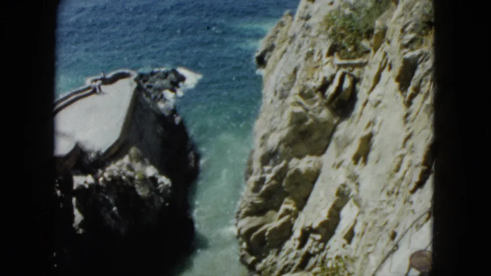 8mm home movie from Acapulco, 1962: cliff face with turquoise surf crashing at base, sparse vegetation and cliff-top structure visible mid-frame.