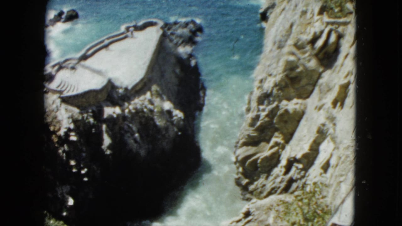 8mm home-movie frame circa 1962 showing Acapulco coastal cliffs, a concrete viewing platform on a rocky outcrop, and turquoise Pacific water.