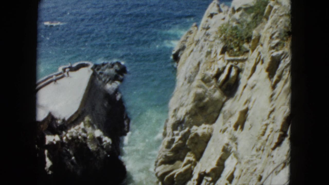 8mm frame circa 1962 showing Acapulco cliff face mid-pan, with concrete platform and railings visible on rocky outcrop above turquoise sea.