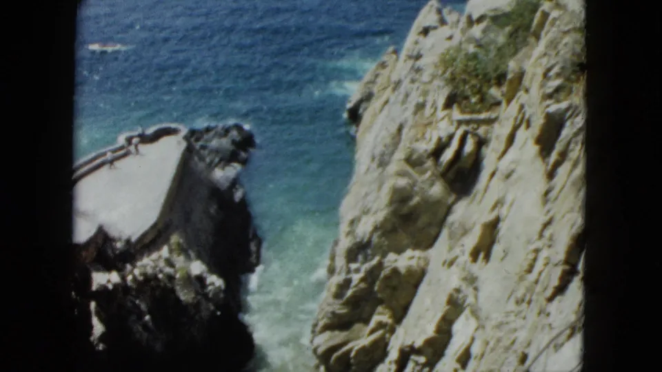 8mm frame circa 1962 showing Acapulco cliff face mid-pan, with concrete platform and railings visible on rocky outcrop above turquoise sea.
