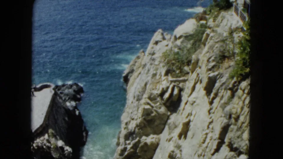 8mm home-movie frame circa 1962 showing Acapulco cliff face close-up with dark scrubby vegetation and deep blue Pacific water.
