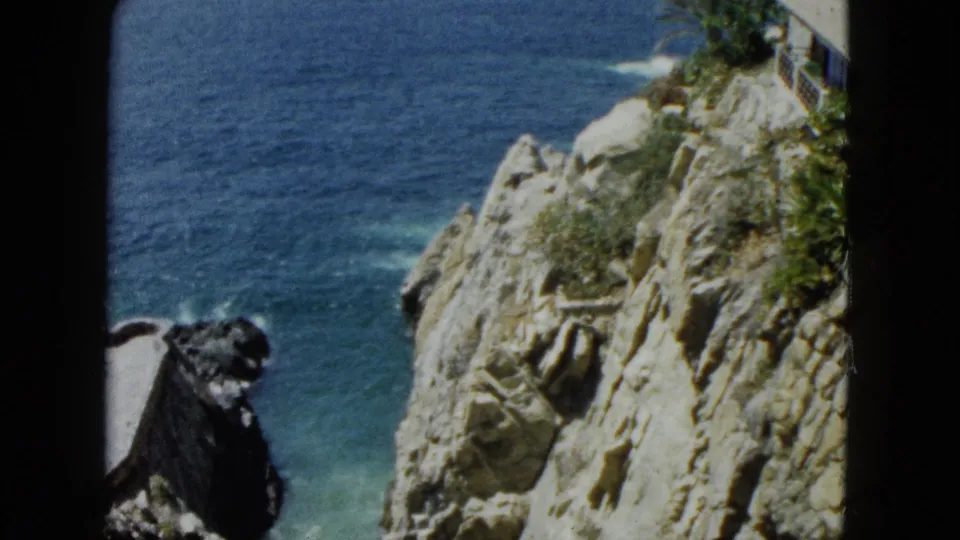 8mm home-movie frame circa 1962, Acapulco coastal cliffs with scrubby vegetation and blue Pacific behind jagged rock face.
