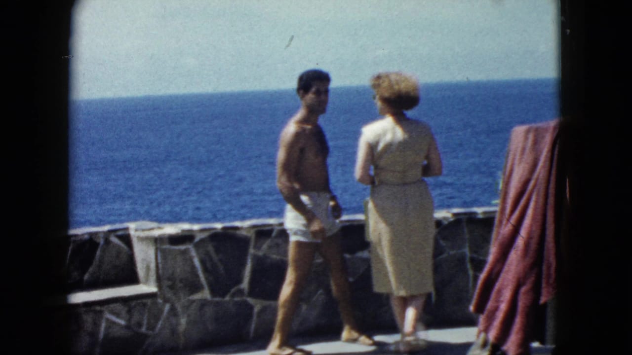 Couple pausing on an oceanside stone terrace, man in partial turn toward woman, red draped cloth at right, Acapulco area, circa 1962.