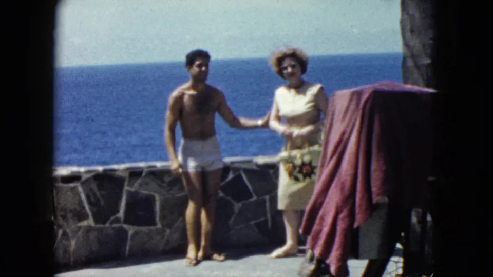 Couple in face-to-face conversation on ocean terrace, woman with hat and floral accessory, red draped cloth in background, Acapulco area, circa 1962.