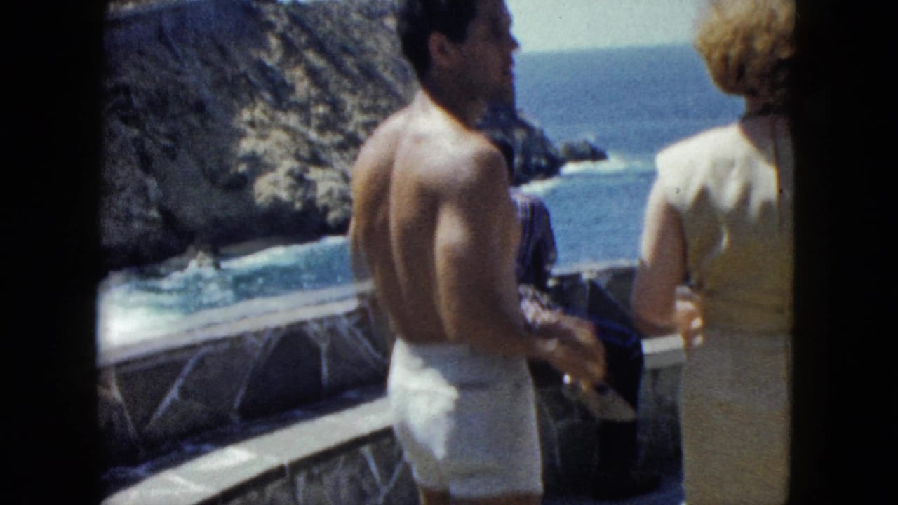 Shirtless man in white swim trunks and woman in cream shift dress on a stone terrace overlooking the Pacific coast, Acapulco, circa 1962, 8mm film.