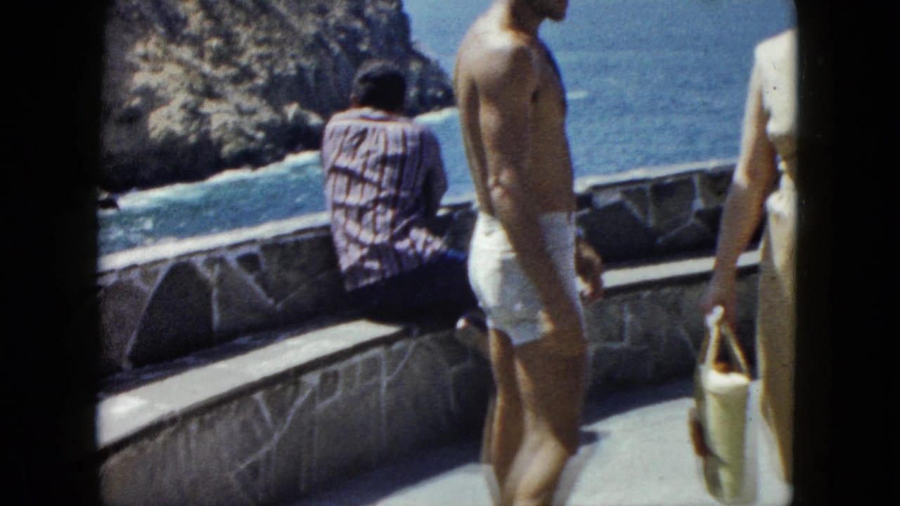 Three figures on a stone terrace wall, one seated in a plaid shirt, rocky cliffs and ocean visible beyond.