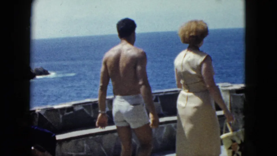 Man in white swim trunks and woman in cream dress walk a stone terrace above the Pacific, Acapulco, circa 1962, 8mm home movie.