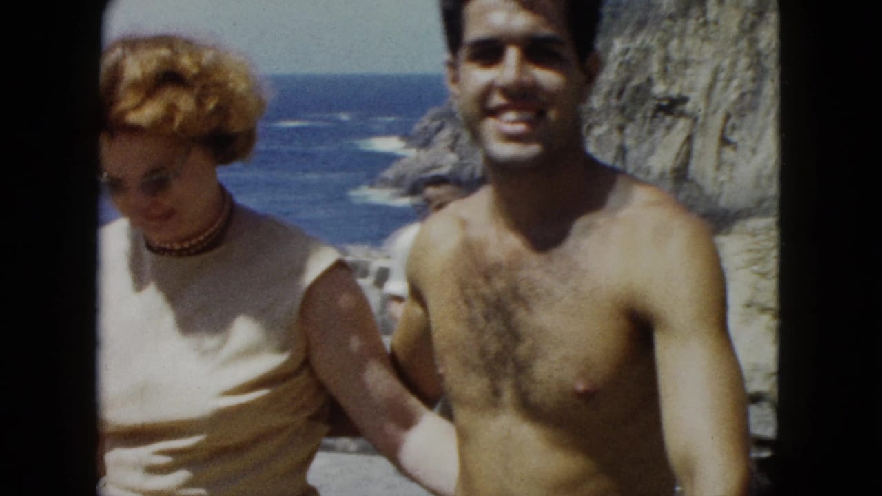 Young couple at a coastal overlook, likely Acapulco, Mexico, circa 1962; shirtless man smiling toward camera, woman with auburn curly hair and bead necklace, rocky Pacific cliffs in background.