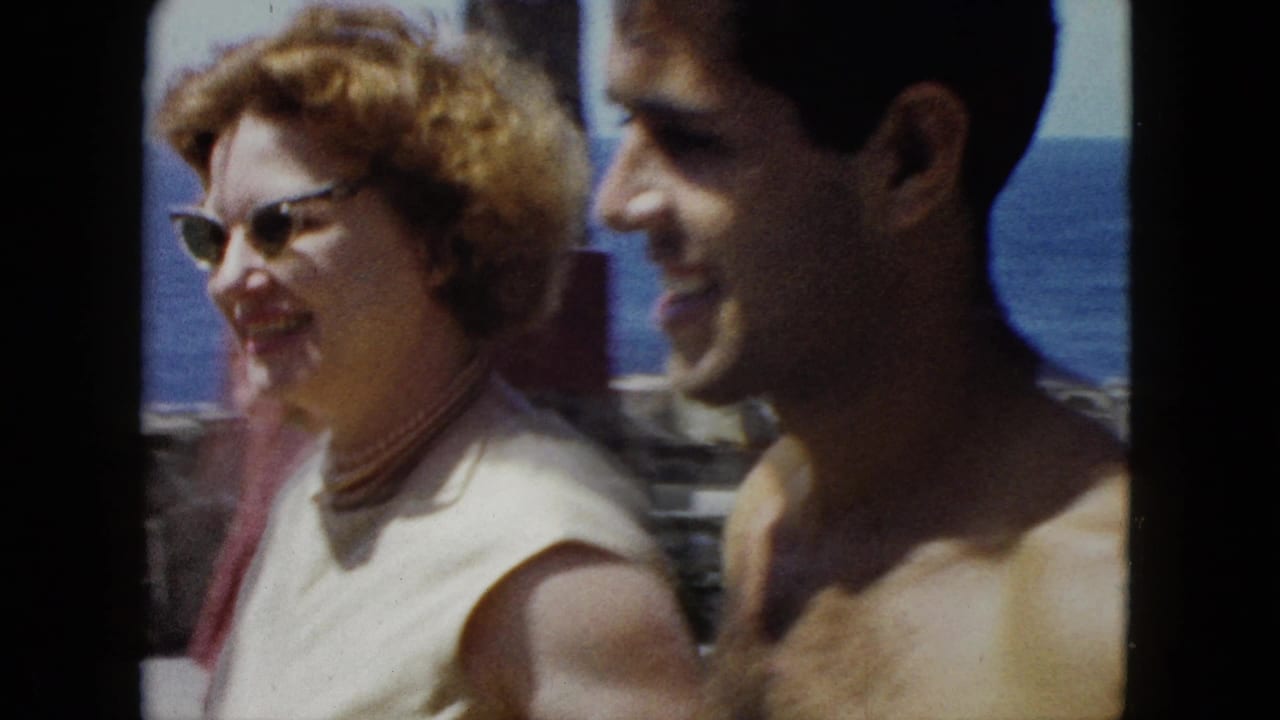 Close-up of a woman in cat-eye sunglasses laughing beside a shirtless smiling man; rocky coastline and blue ocean visible behind them.