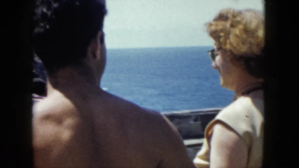 Shot from behind the couple at what may be a coastal overlook in Acapulco, circa 1962; woman with cat-eye sunglasses faces the Pacific, rocky horizon line visible under a clear sky.