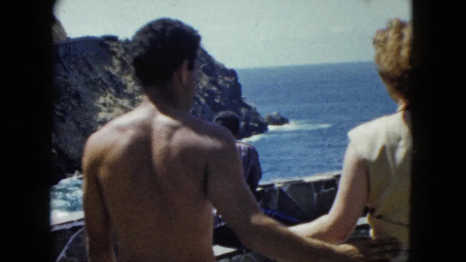Couple facing rocky Pacific cliffs at a coastal overlook, possibly Acapulco, circa 1962; a third figure may be partially visible at the far right edge of frame.