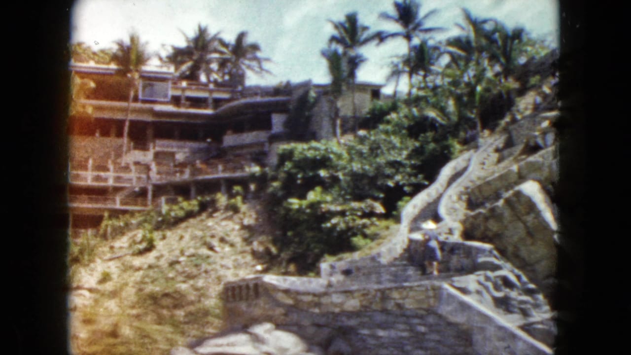 Stone staircases winding up a rocky hillside with palm trees and a multi-story building above; one figure visible on the steps.
