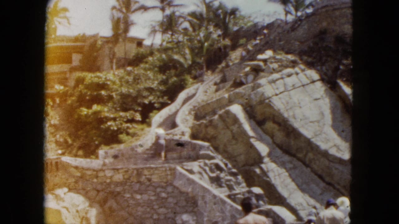 Stone stairs and large boulders on a sunlit hillside with palm trees above, a small figure at lower right, and a building in the background.