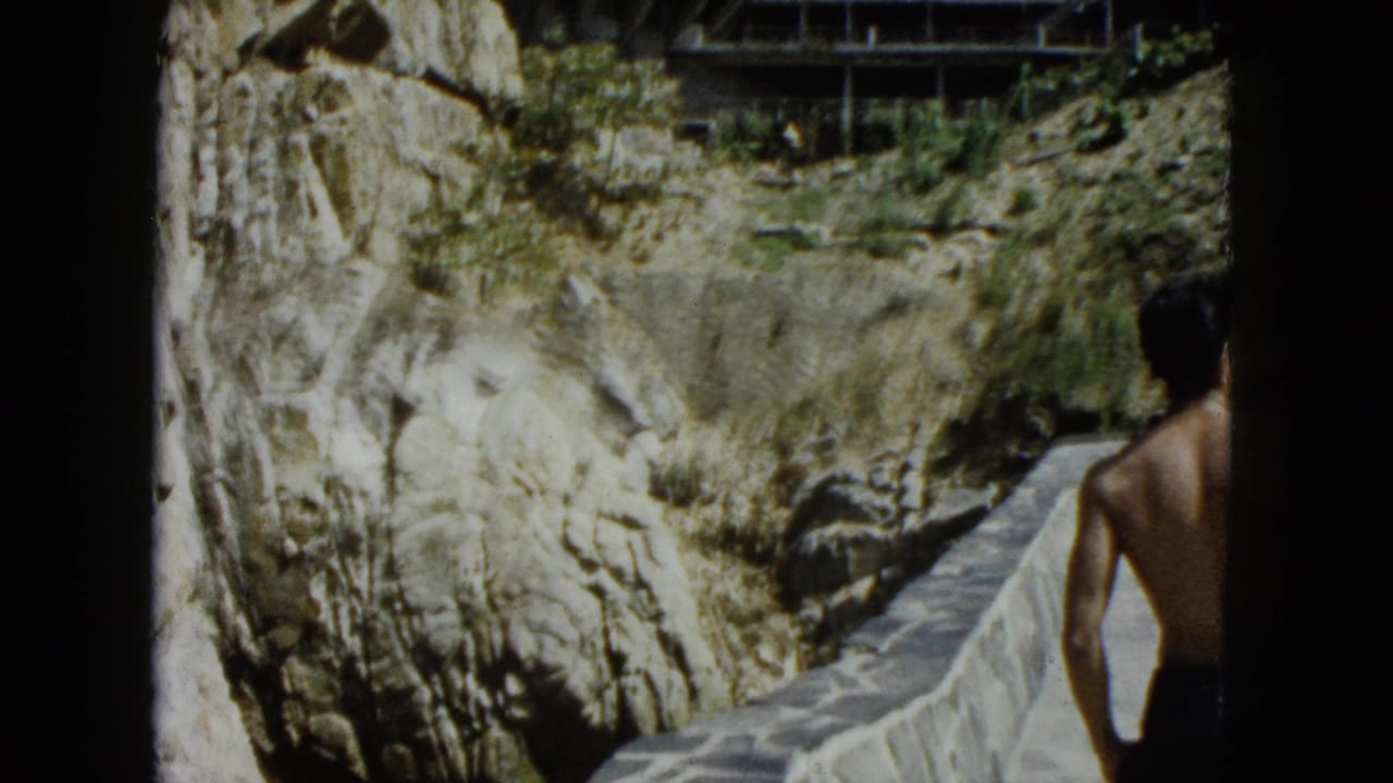 Shirtless man viewed from behind on a stone path beside a rugged cliff face, 8mm home movie.