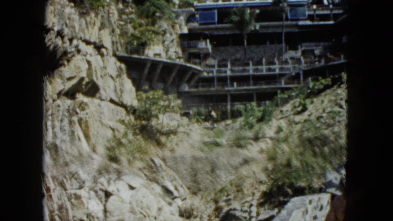 Multi-tiered building with open balconies built against a rocky cliff face, 8mm film.