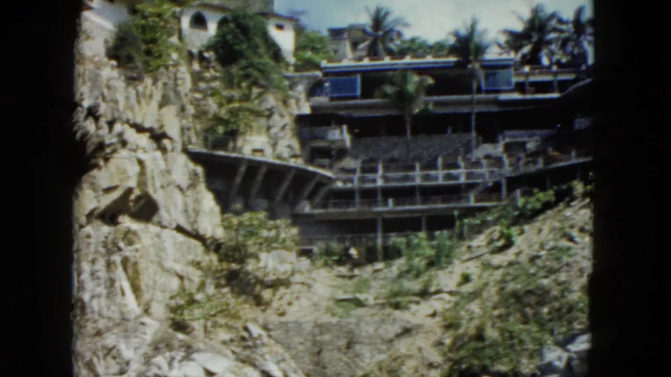 Multi-story cliffside resort hotel with tiered open balconies, palm trees, and blue architectural accents, Acapulco, Mexico, circa 1962, 8mm film.