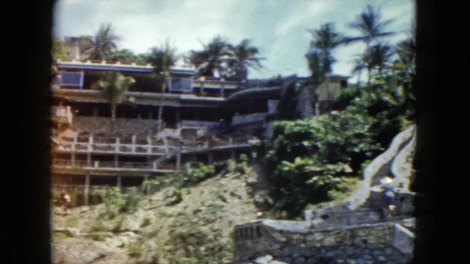 Wide view of multi-tiered cliffside resort with palm trees and blue sky, Acapulco, Mexico, circa 1962; possible small figures visible at lower right.