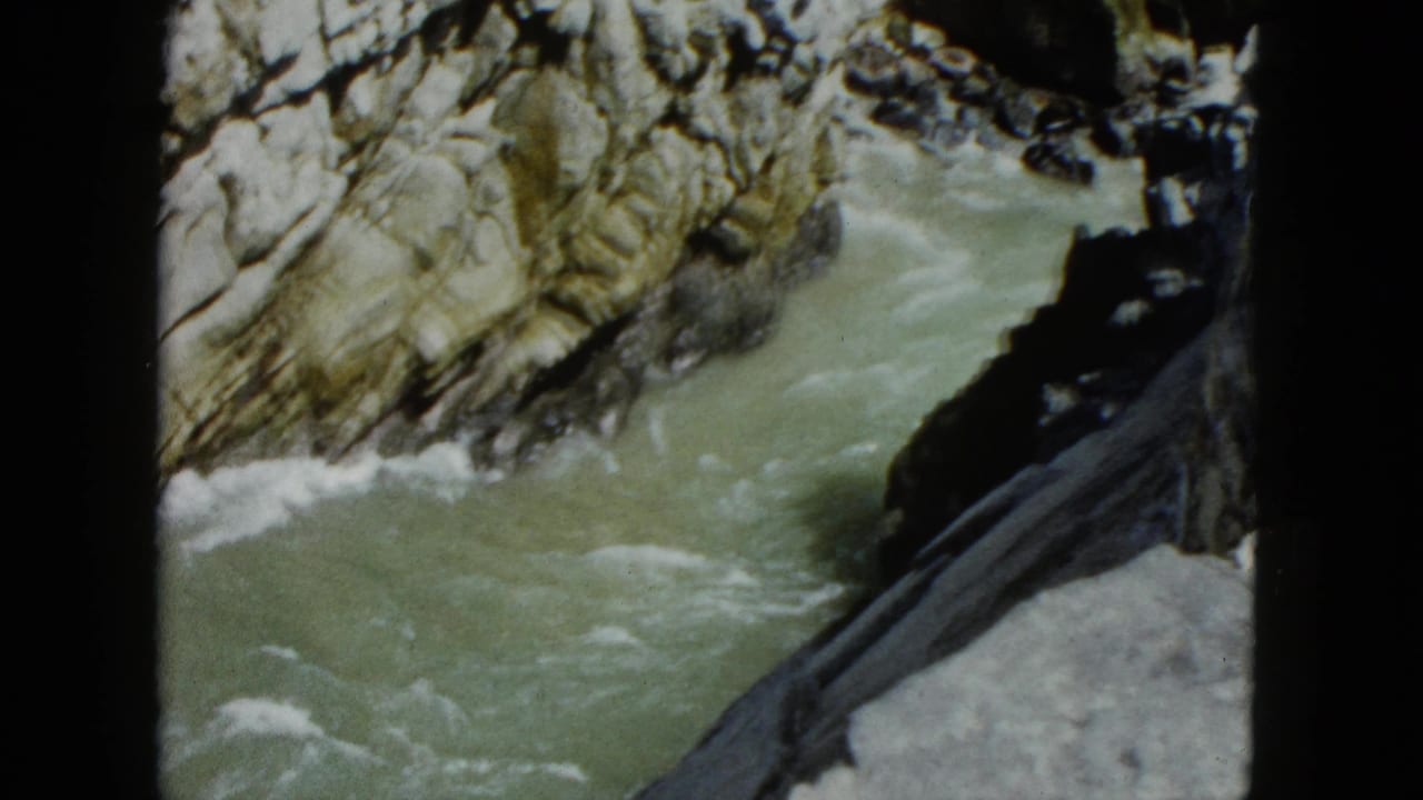 Turbulent green water rushing through a sunlit rocky canyon, white foam against cliff faces, filmed from above.