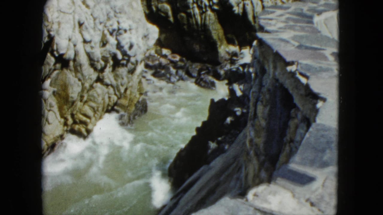 8mm frame showing coastal rocky cliffs with crashing white foam and a stone path at lower right, Acapulco area, circa 1962.