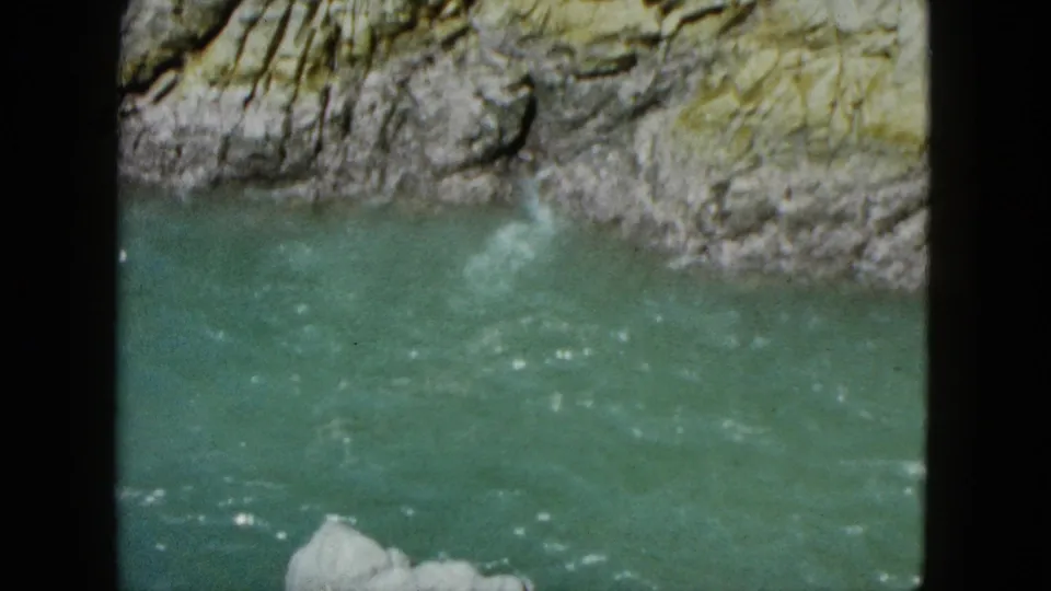 1962 Acapulco home-movie frame showing green-blue river water moving below a layered rock face, with dark film-frame edges visible.