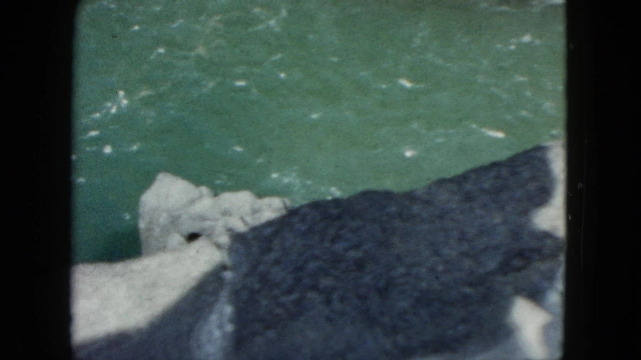 1962 Acapulco frame with pale river boulders, green moving water, and a large dark rock or shadow crossing the foreground.
