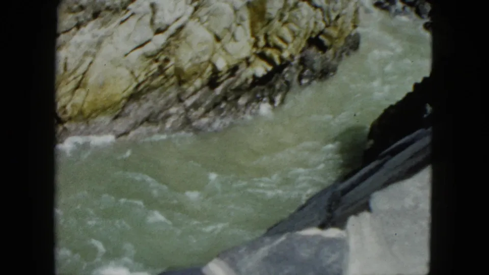 1962 Acapulco frame showing greenish river water flowing past jagged layered rocks, partly blocked by a dark foreground edge.